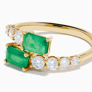 The 14K Yellow Gold Diamond and Emerald Bypass Ring features two rectangular green emeralds and rows of sparkling round white diamonds on each side in an open band design, set against a white background.