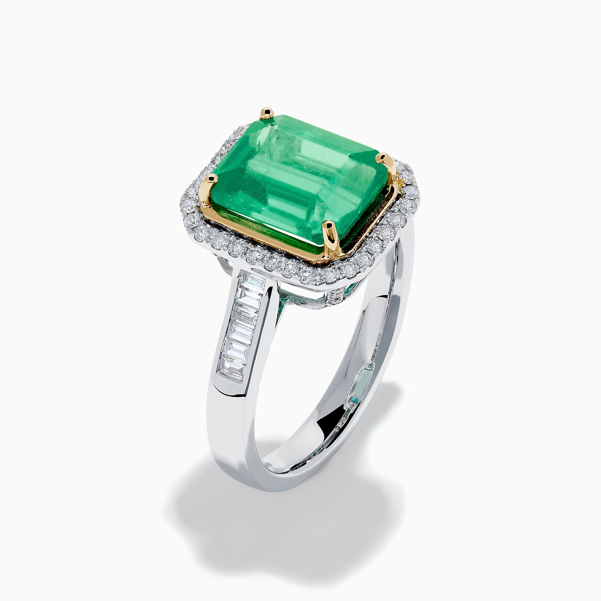 The Brasilica 14K Two Tone Gold Emerald and Diamond Ring showcases a large rectangular green emerald in a gold prong setting, accented by a halo of clear diamonds and baguette-cut diamonds along the band.