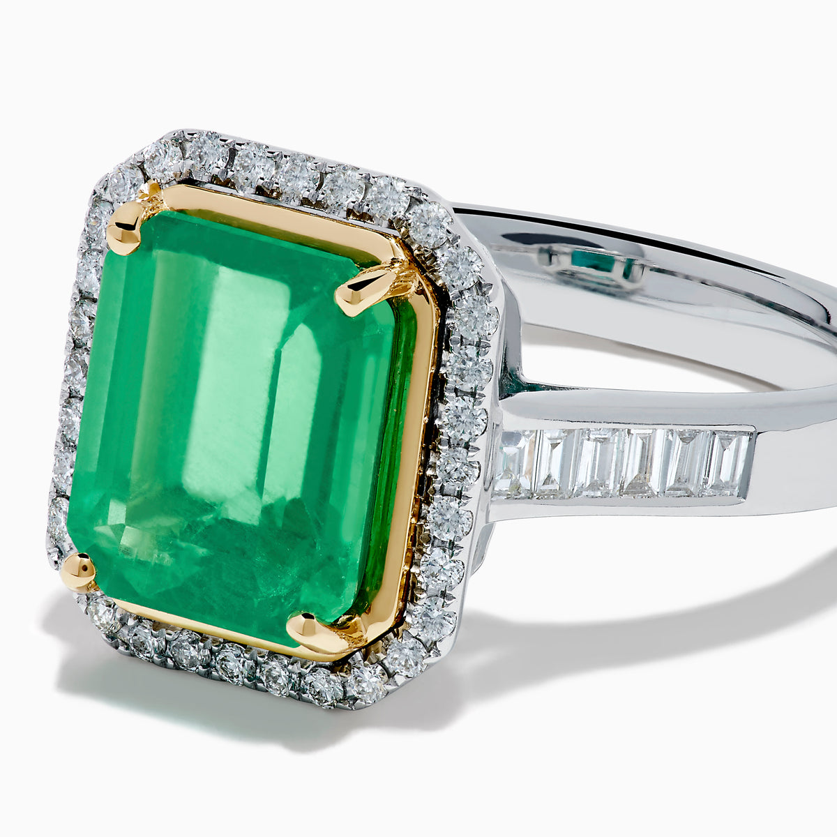 The Brasilica 14K Two Tone Gold Emerald and Diamond Ring showcases a large rectangular green emerald in a gold prong setting, accented by a halo of clear diamonds and baguette-cut diamonds along the band.