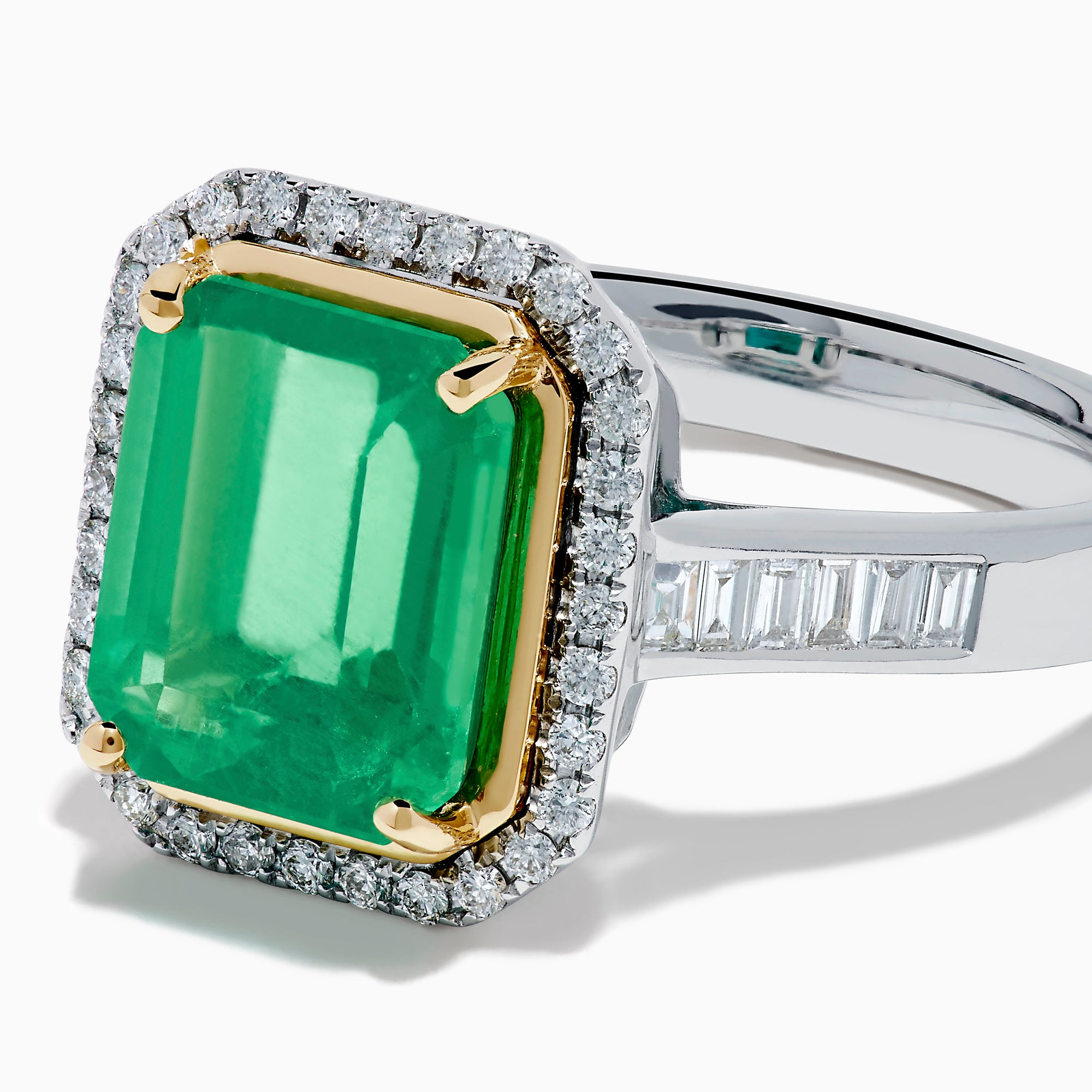 The Brasilica 14K Two Tone Gold Emerald and Diamond Ring showcases a large rectangular green emerald in a gold prong setting, accented by a halo of clear diamonds and baguette-cut diamonds along the band.