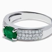 The Brasilica 14K White Gold Emerald and Diamond Ring showcases a central oval green emerald with three rows of round diamonds on each side, set in white gold and shown against a white background.