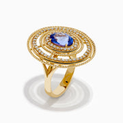 The 14K Yellow Gold Tanzanite and Diamond Concentric Halo Ring showcases concentric circular diamond bands with a central oval tanzanite gemstone, elegantly presented on a white background.