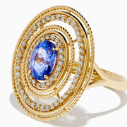 The 14K Yellow Gold Tanzanite and Diamond Concentric Halo Ring showcases concentric circular diamond bands with a central oval tanzanite gemstone, elegantly presented on a white background.