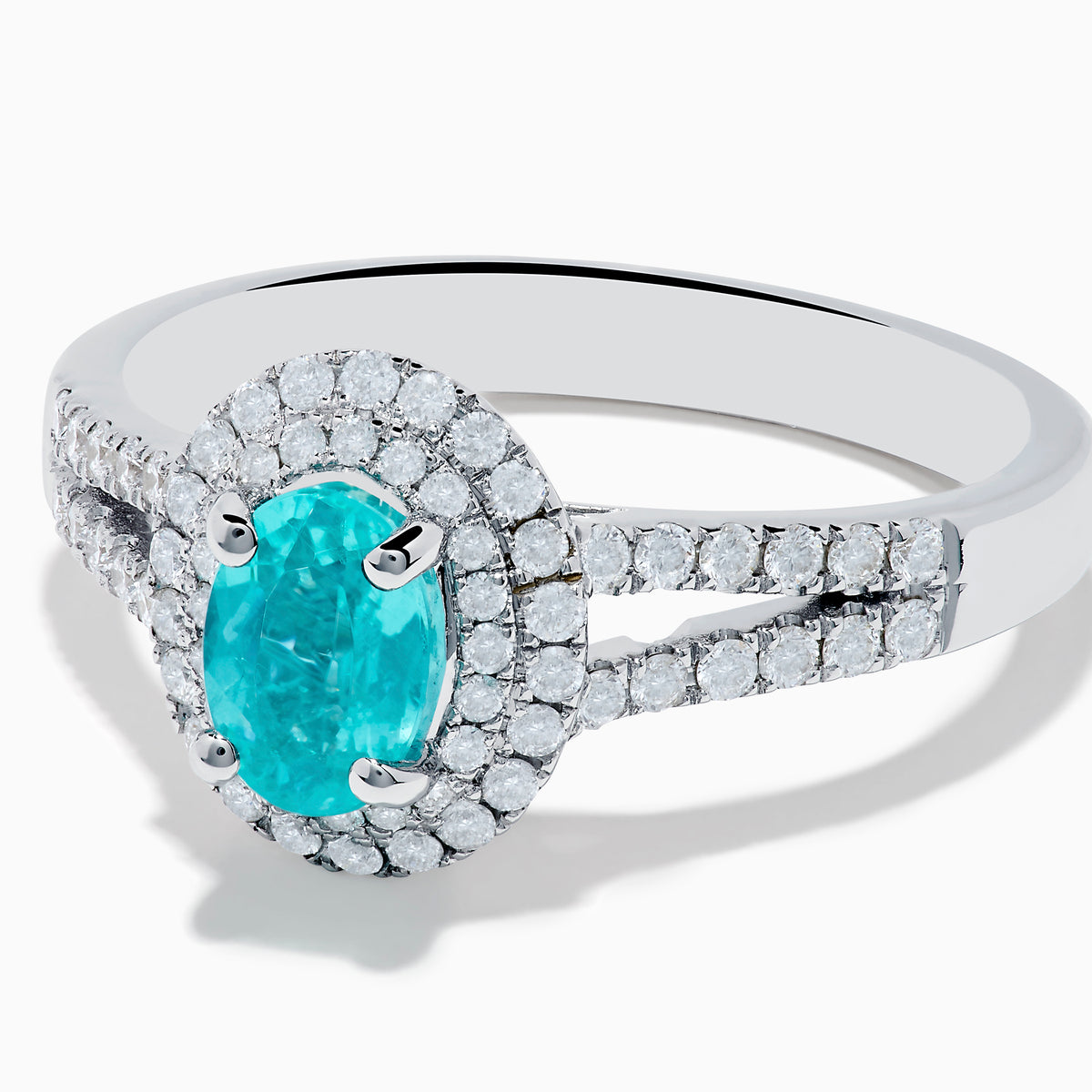 The 18K White Gold Paraiba and Diamond Ring showcases an oval Paraiba gemstone at the center, encircled by a halo of sparkling diamonds and accented with additional diamonds along its elegant split shank band.