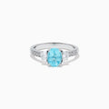 The 14K White Gold Paraiba and Diamond Three Stone Ring features an oval blue Paraiba at the center, flanked by two diamonds, with additional diamonds set along each side of the band.