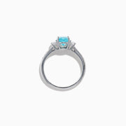 The 14K White Gold Paraiba and Diamond Three Stone Ring features an oval blue Paraiba at the center, flanked by two diamonds, with additional diamonds set along each side of the band.