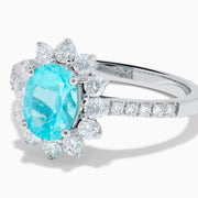 The 14K White Gold Paraiba and Diamond Halo Ring showcases an oval-shaped Paraiba gemstone at the center, encircled by a sparkling diamond halo with additional diamonds adorning the band.