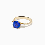 The Nahla Siri 14K Yellow Gold Tanzanite and Diamond Ring showcases a large cushion-cut tanzanite center stone, accented by two tapered white diamonds, displayed on a white background.