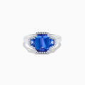 The Nahla 14K White Gold 3 Stone Tanzanite and Diamond Ring features an emerald-cut tanzanite center flanked by sparkling diamonds, all set in a geometric design and displayed on a white background.