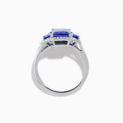 The Nahla 14K White Gold 3 Stone Tanzanite and Diamond Ring features an emerald-cut tanzanite center flanked by sparkling diamonds, all set in a geometric design and displayed on a white background.