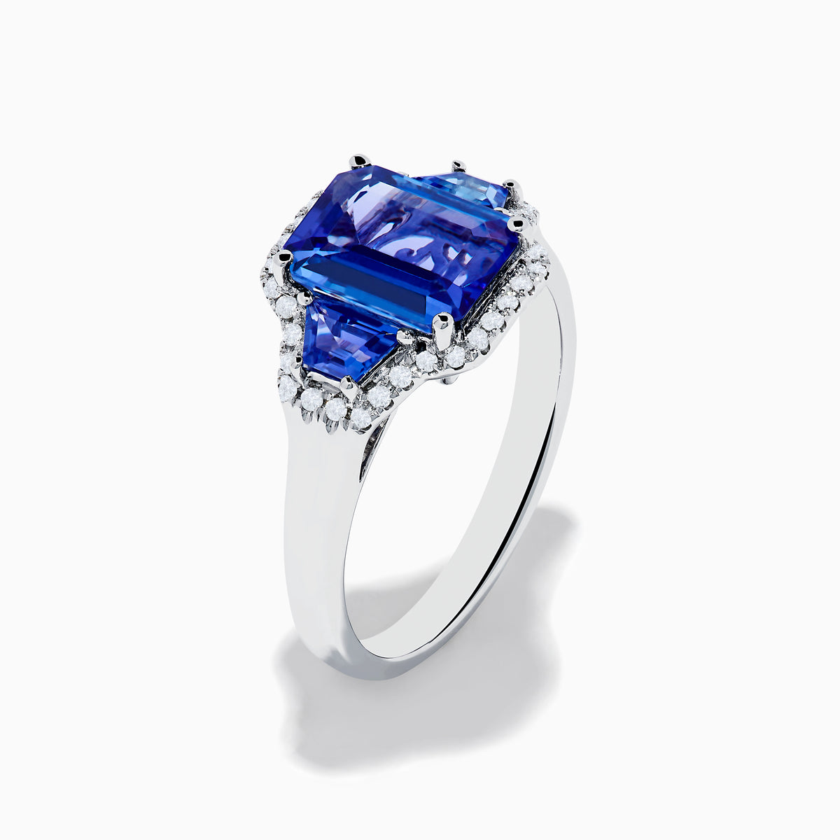 The Nahla 14K White Gold 3 Stone Tanzanite and Diamond Ring features an emerald-cut tanzanite center flanked by sparkling diamonds, all set in a geometric design and displayed on a white background.