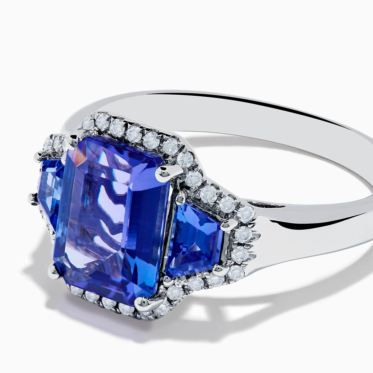 The Nahla 14K White Gold 3 Stone Tanzanite and Diamond Ring features an emerald-cut tanzanite center flanked by sparkling diamonds, all set in a geometric design and displayed on a white background.