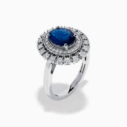The 14K White Gold Blue Sapphire and Diamond Double Halo Ring features an oval blue sapphire surrounded by two halos of round white diamonds on a white gold band, shown against a white background.