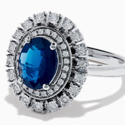 The 14K White Gold Blue Sapphire and Diamond Double Halo Ring features an oval blue sapphire surrounded by two halos of round white diamonds on a white gold band, shown against a white background.