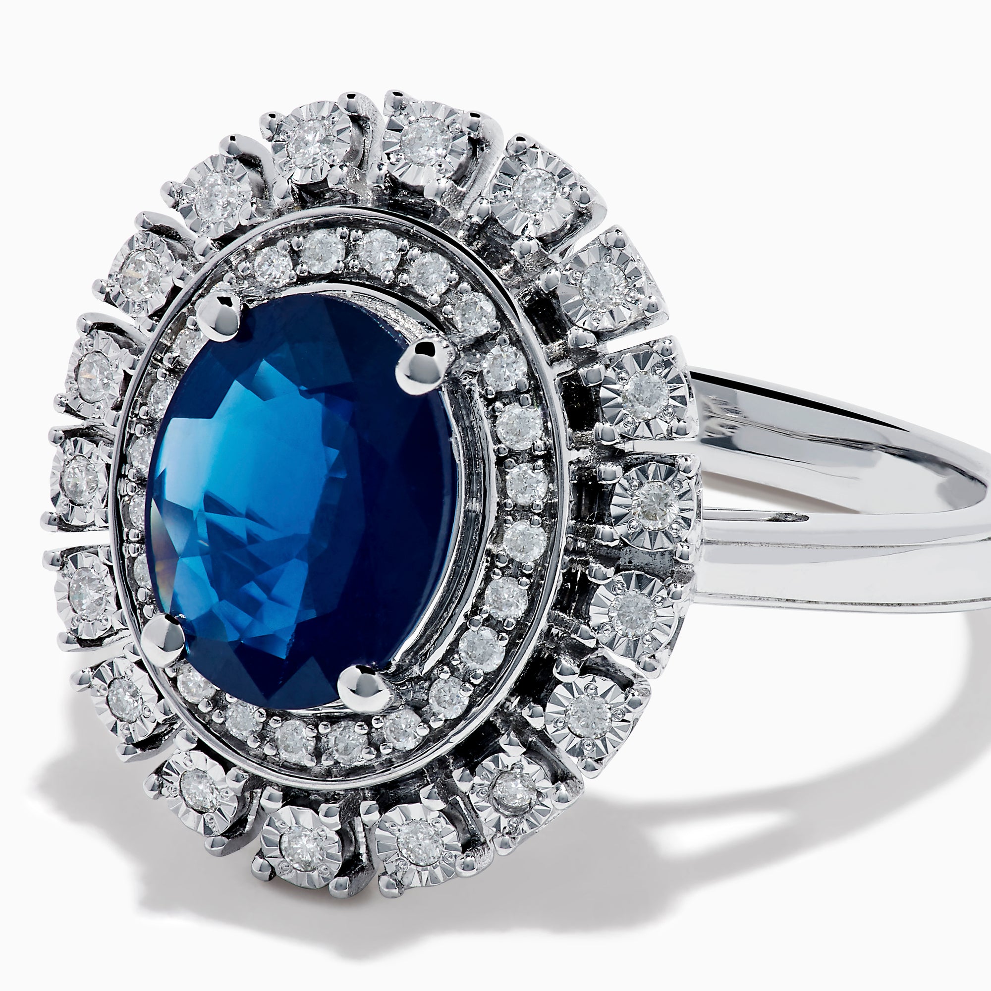 The 14K White Gold Blue Sapphire and Diamond Double Halo Ring features an oval blue sapphire surrounded by two halos of round white diamonds on a white gold band, shown against a white background.