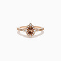 The 14K Rose Gold Brown and White Diamond Halo Ring showcases an oval brown and white diamond center, encircled by a halo of white diamonds and set with additional diamonds along the band.
