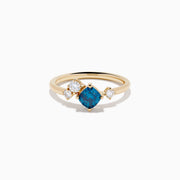 The 14K Yellow Gold Blue and White Diamond Ring showcases a cushion-cut blue stone center with two round white diamonds on each side, set against a white background.