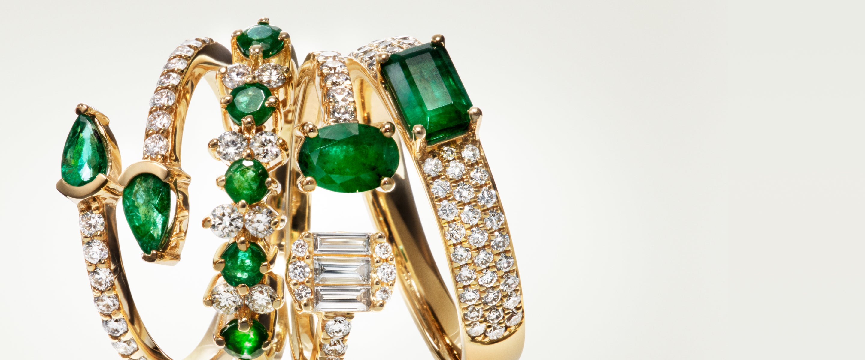 Four gold rings adorned with sparkling diamonds and vibrant green emeralds, featuring various shapes and arrangements, displayed against a plain light background.