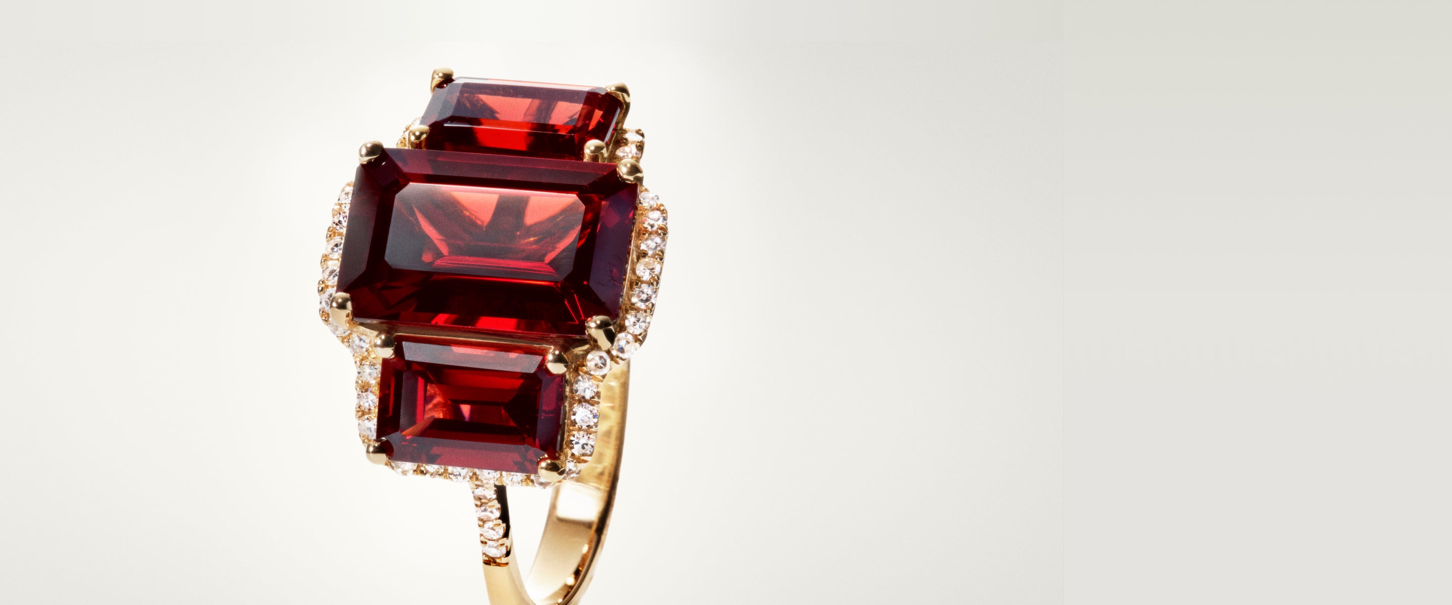 A gold ring featuring three large rectangular red gemstones arranged vertically, each bordered by small round diamonds, displayed against a light, neutral background.