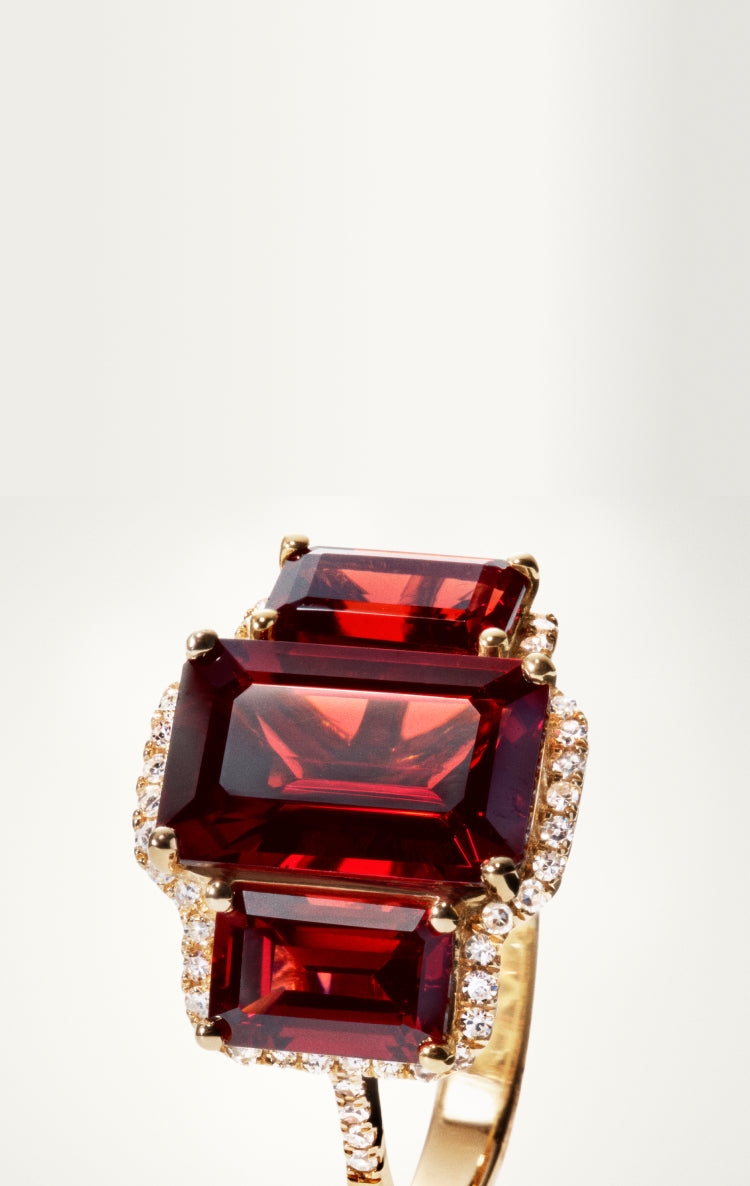 A gold ring featuring three large, rectangular red gemstones arranged vertically, each surrounded by small clear diamonds, set against a plain white background.