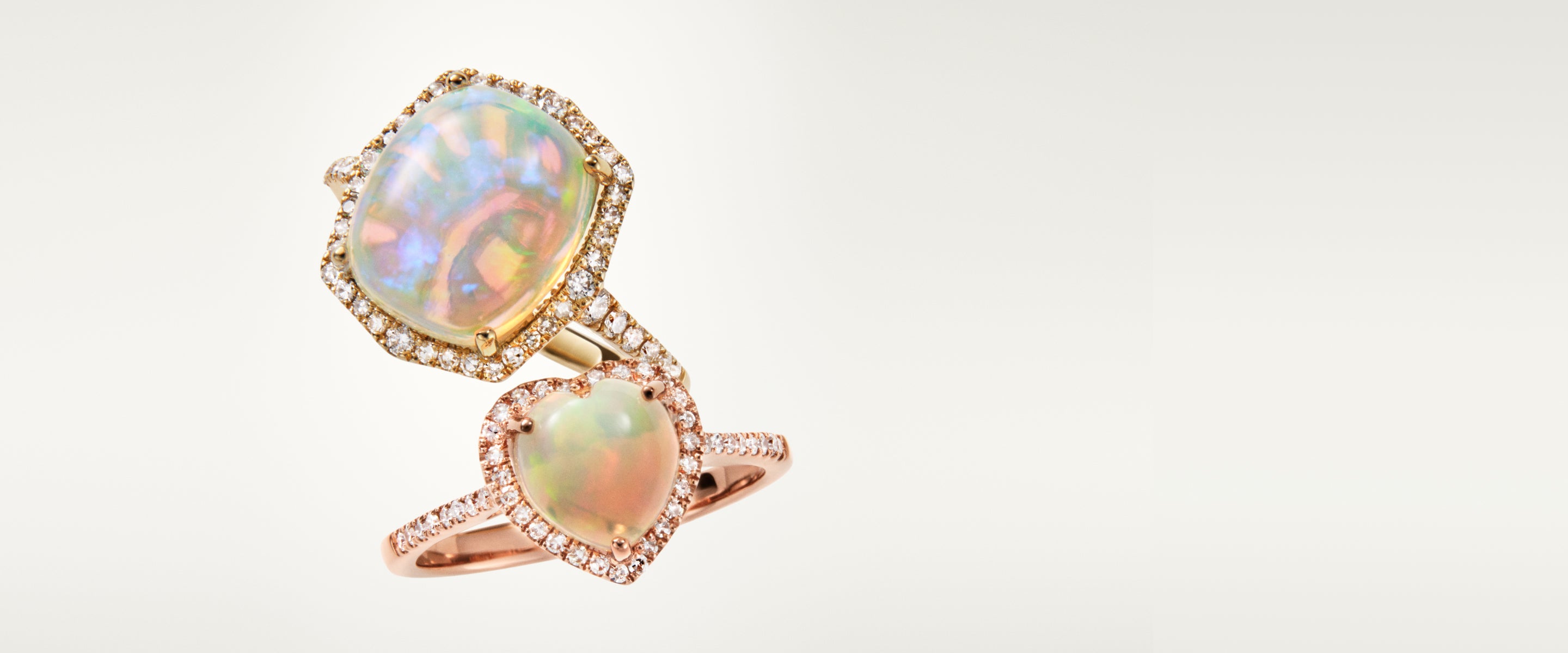 A gold ring featuring two opal gemstones, one oval and one heart-shaped, each surrounded by small diamonds, set against a plain light background.