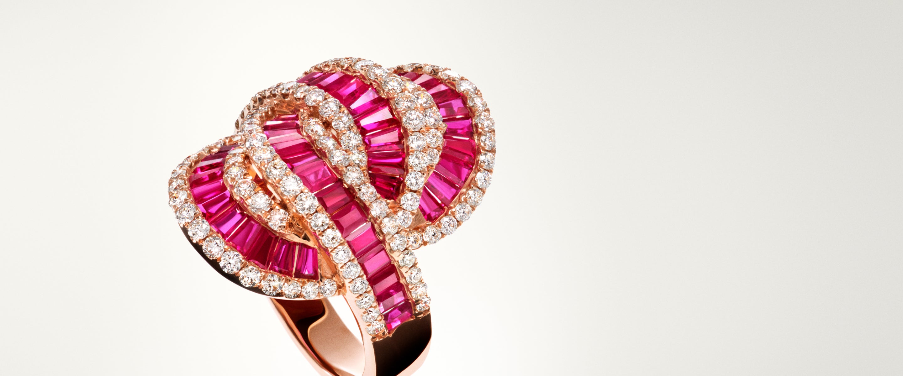A gold ring featuring an intricate, overlapping design with vivid pink gemstones set in rows, bordered by sparkling white diamonds against a plain light background.