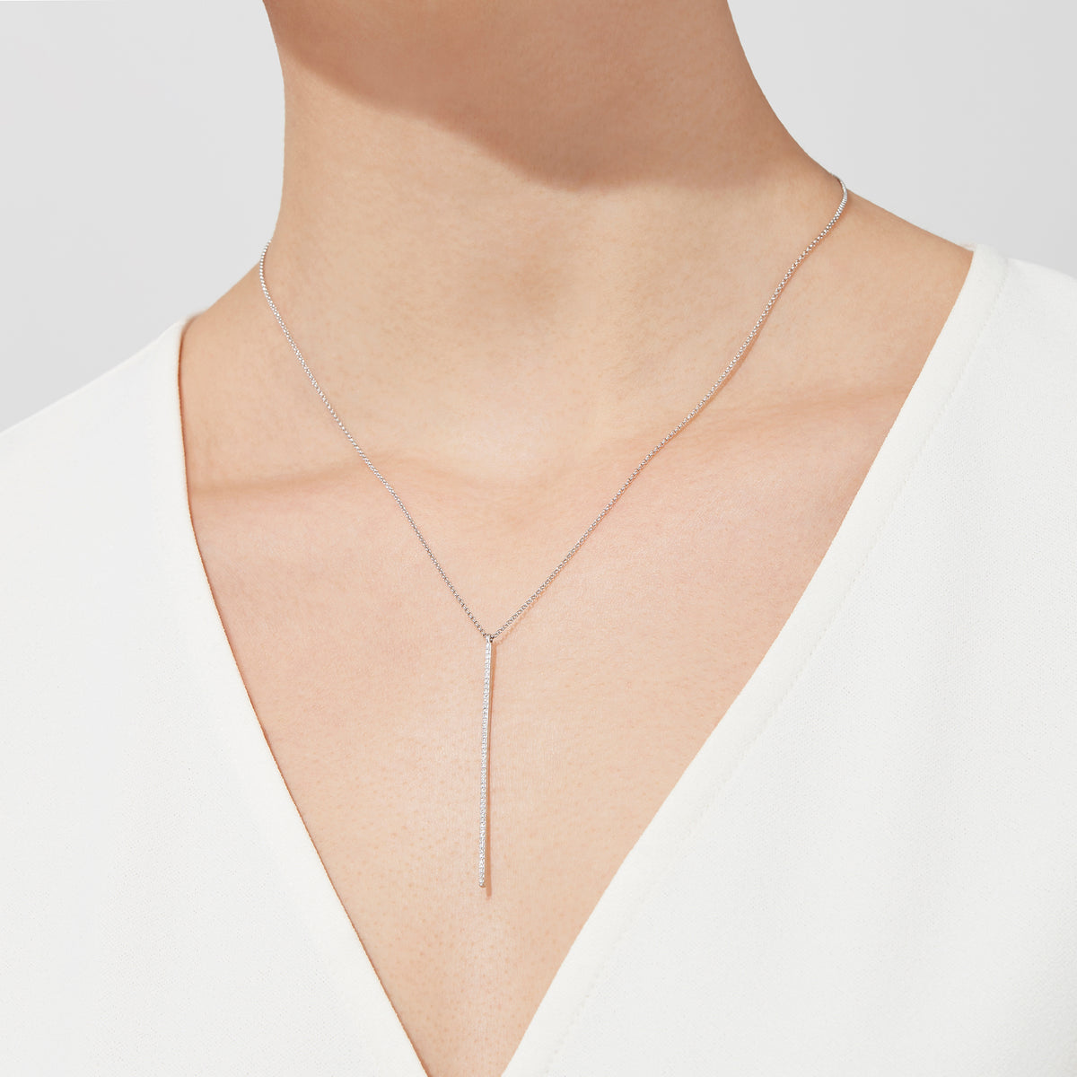 A person in a white V-neck top wears the Pave Classica 14K White Gold Diamond Vertical Pendant (0.14 TCW), featuring a delicate lariat style, against a plain light background.