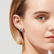 Close-up of a woman with short, light brown hair and green eyes wearing Ocean Bleu 14K Yellow Gold Blue Topaz and Diamond Earrings, 9.70 TCW. The plain background highlights her fair skin and calm, elegant look.
