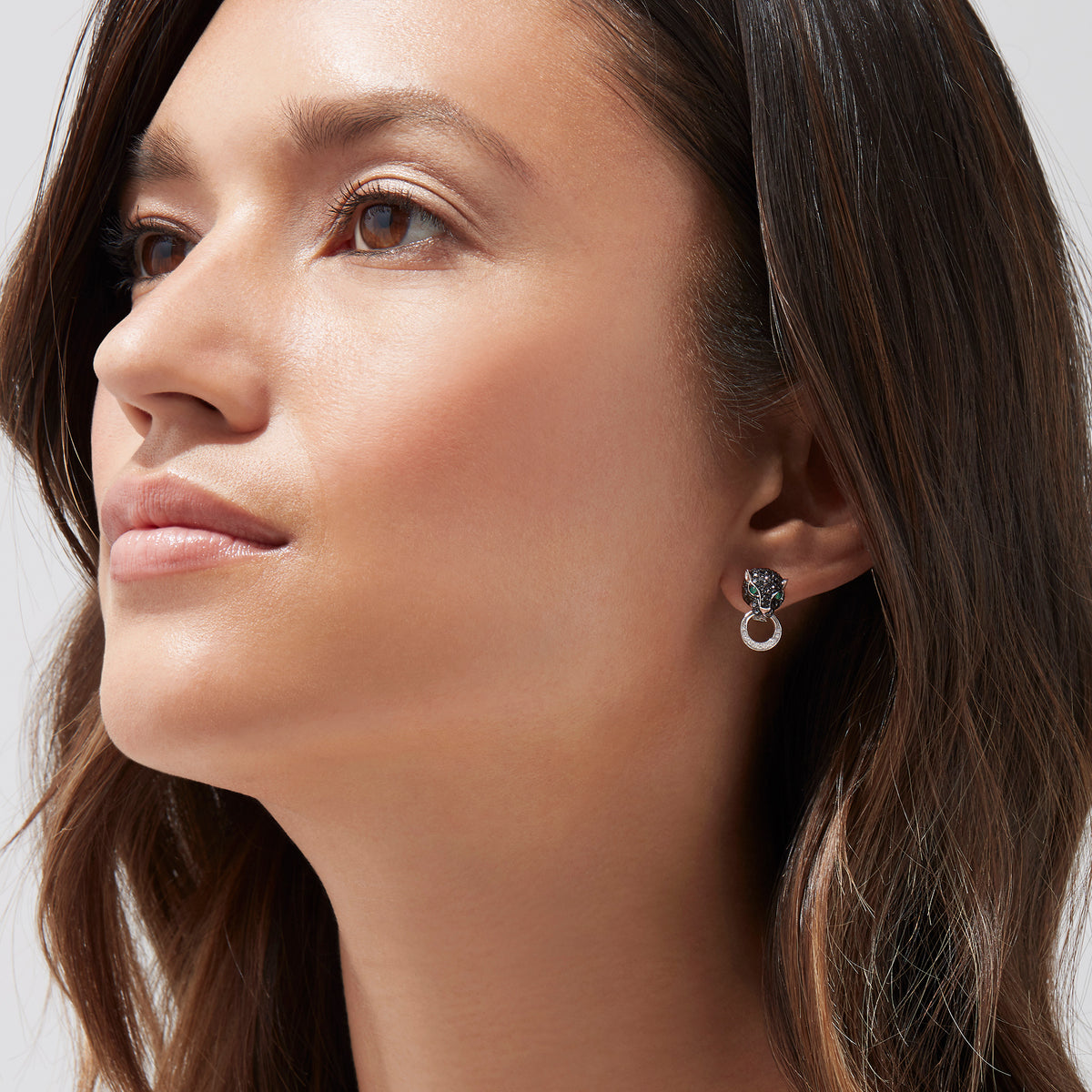 A woman with long brown hair looks upwards, wearing Signature 14K White Gold Black Diamond & Emerald Earrings (0.84 TCW) with a circular, textured design, set against a plain light background.