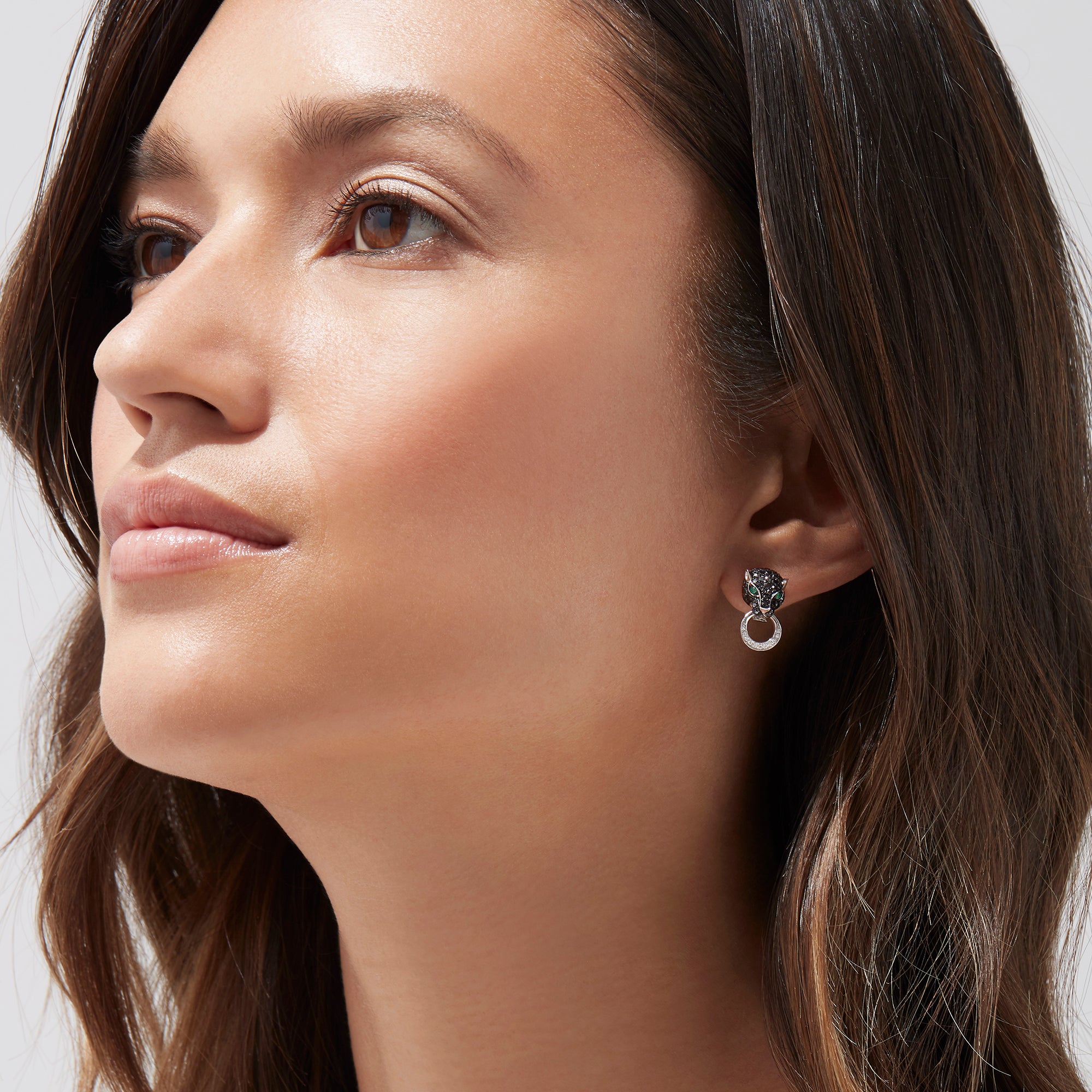 A woman with long brown hair looks upwards, wearing Signature 14K White Gold Black Diamond & Emerald Earrings (0.84 TCW) with a circular, textured design, set against a plain light background.