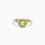 The 14k Yellow Gold Peridot and Diamond Ring, 1.10 TCW, showcases a round green peridot center encircled by a halo of sparkling diamonds, all set in yellow gold and displayed against a white background.