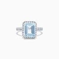 The Aquarius 14K White Gold Aquamarine and Diamond Ring, 3.33 TCW, features a large emerald-cut aquamarine surrounded by a diamond halo and set on a diamond-studded band, shown on a white background.