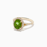 The 14K Yellow Gold Peridot and Diamond Ring showcases an oval-cut green peridot center, encircled by a halo of round white diamonds, with extra diamonds set along the band.