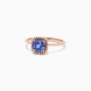 The Nahla Siri 14K Rose Gold Tanzanite and Diamond Ring (0.81 TCW) showcases a cushion-cut blue tanzanite encircled by a halo of clear diamonds, set against a white background.